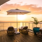 A collection of rattan lounge chairs, a large umbrella and a large potted plant face the ocean as the sun sets on the sky deck area at the S Hotel in Jamaica.